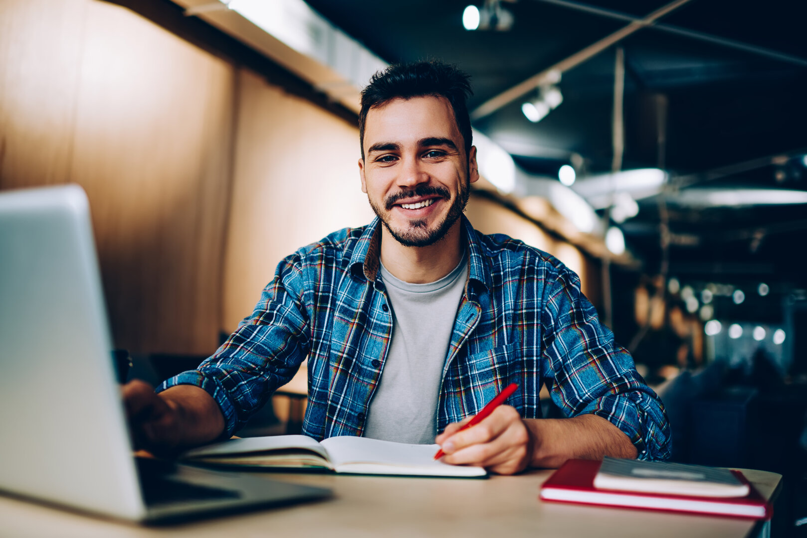 joven estudiante sonriente escribiendo en un cuaderno dentro de una biblioteca moderna