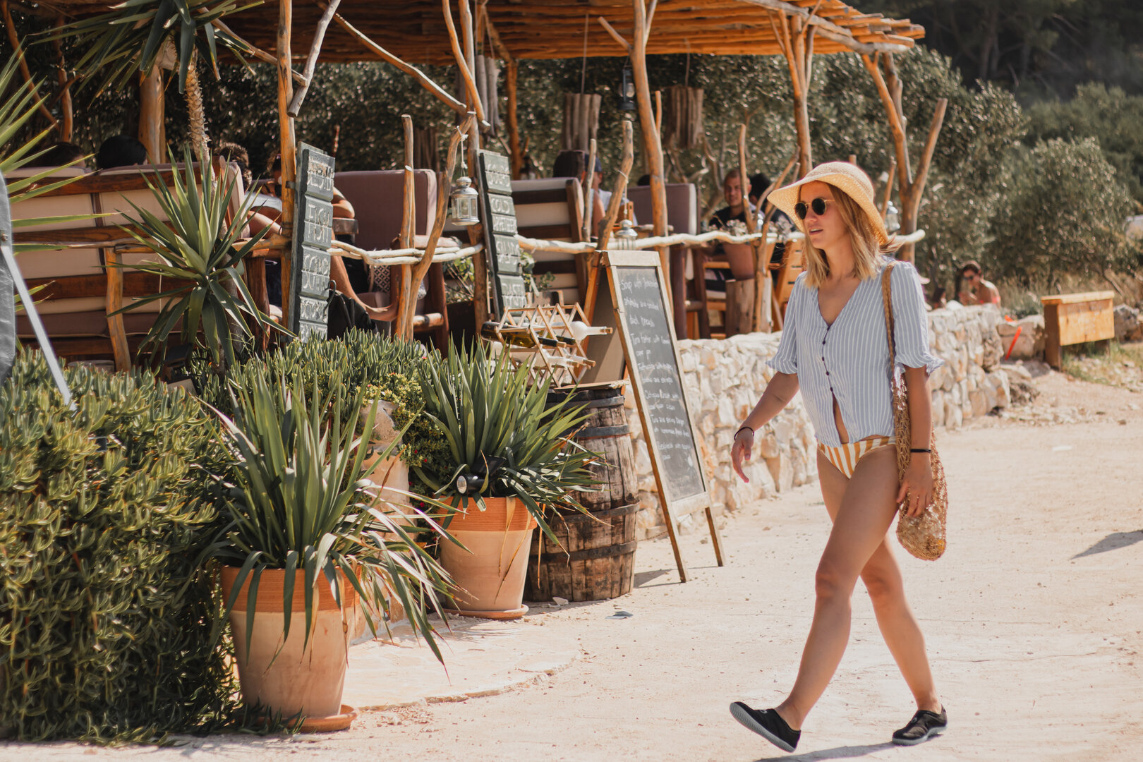 mujer con sombrero de ala ancha caminando junto a una terraza al aire libre en un día soleado